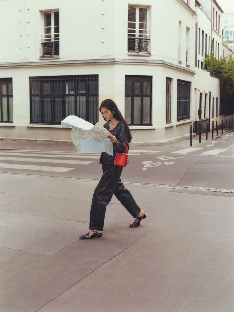 A woman in a black outfit and red purse walks across a crosswalk while holding a city map, with light-colored buildings in the background.