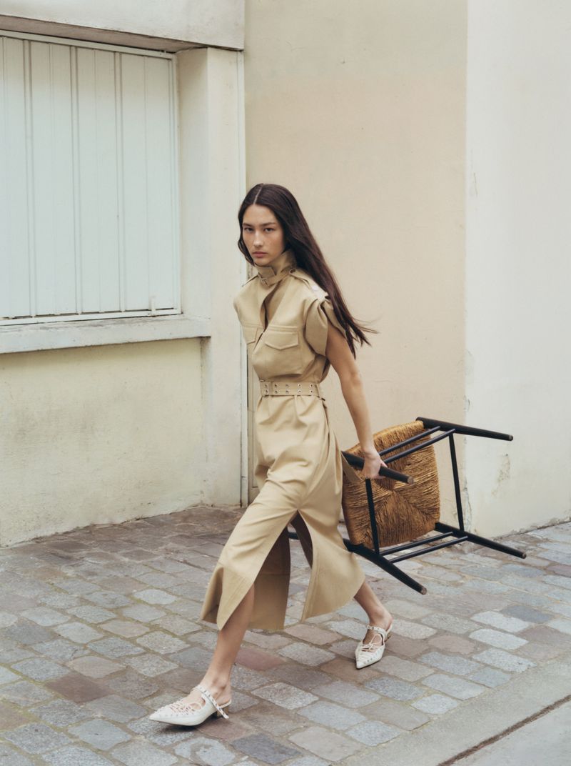 Fashion editorial shot of a woman in a tan dress walking on a cobblestone street carrying a chair, showcasing minimalist style.
