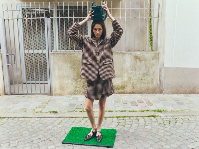 Woman poses outdoors wearing a checkered skirt suit and holding a green purse above her head near a building and metal gate.
