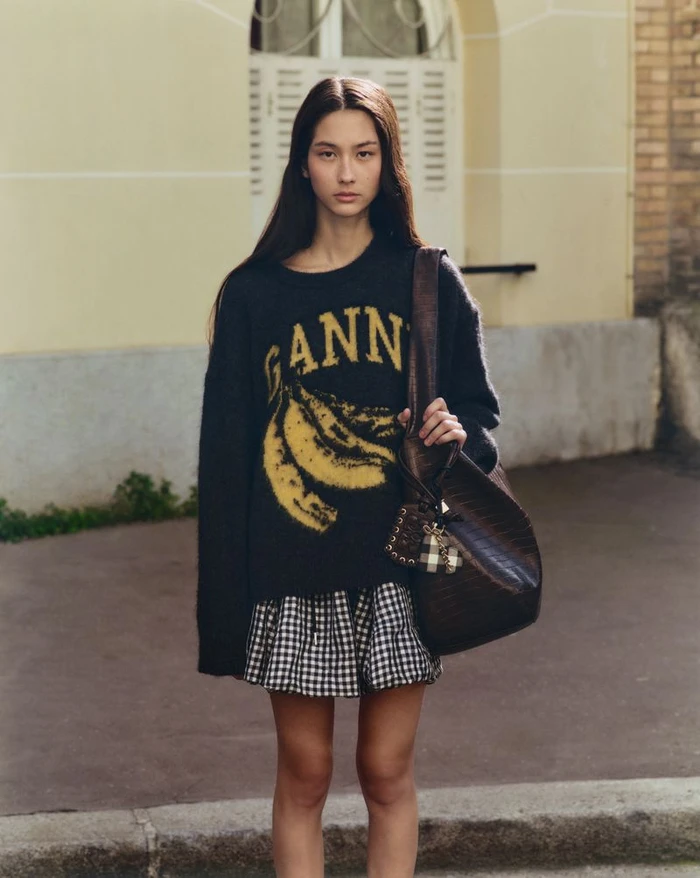 Woman wearing an oversized black GANNI banana sweater, gingham mini skirt, and dark brown shoulder bag on an outdoor street.