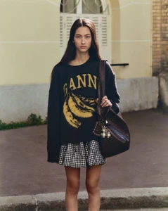 Woman wearing an oversized black GANNI banana sweater, gingham mini skirt, and dark brown shoulder bag on an outdoor street.