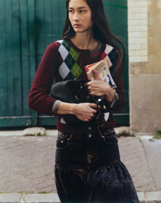 Model carrying a black bag and books, wearing an argyle cardigan and dark denim skirt in front of a green door.