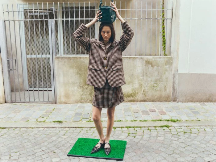 Model wearing a plaid blazer and skirt holding a green handbag on her head while standing on an artificial grass mat outdoors on a stone street.