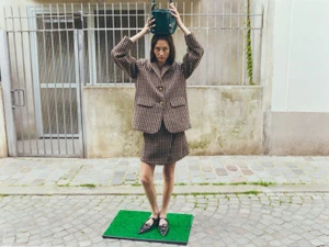 Model wearing a plaid blazer and skirt holding a green handbag on her head while standing on an artificial grass mat outdoors on a stone street.