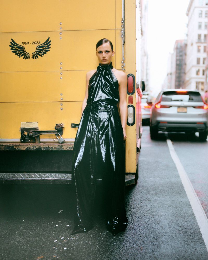 Adult female poses in shiny black halter dress next to yellow truck on city street with blurred cars and buildings in background.