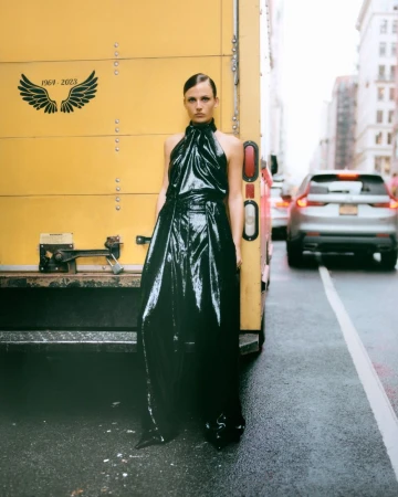 Adult female poses in shiny black halter dress next to yellow truck on city street with blurred cars and buildings in background.