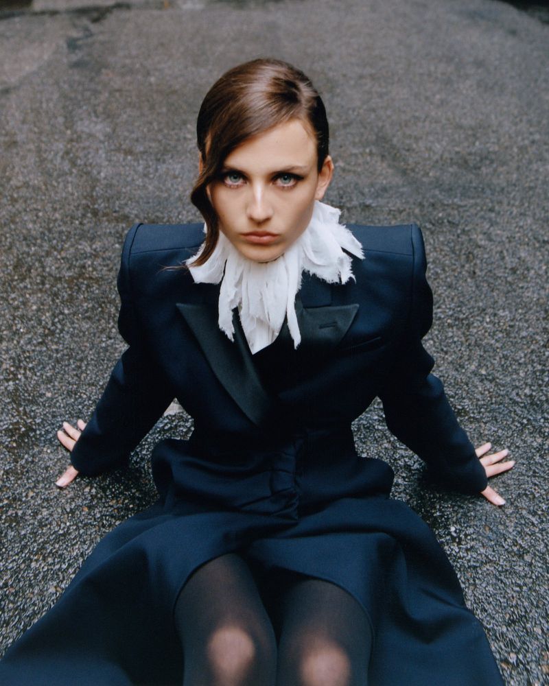Fashion shot of a woman in a dark blazer with a white ruffled collar, sitting on a wet asphalt surface, looking directly at the camera.