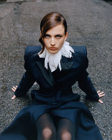 Fashion shot of a woman in a dark blazer with a white ruffled collar, sitting on a wet asphalt surface, looking directly at the camera.