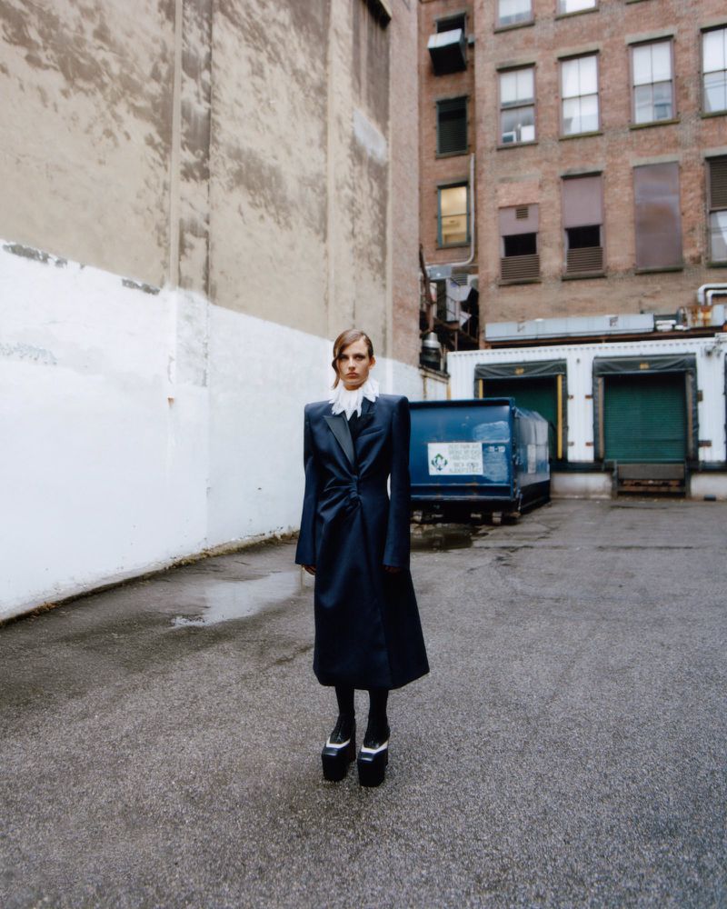 A fashion shot of an adult woman in a long black coat and platform shoes, standing outdoors in front of a brick building in an urban setting.