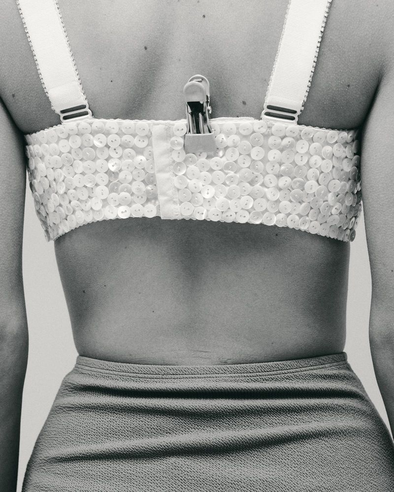 Close-up shot from behind of a person wearing a white beaded bra top and blue skirt. A metal clip is attached to the bra.