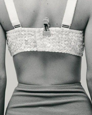 Close-up shot from behind of a person wearing a white beaded bra top and blue skirt. A metal clip is attached to the bra.