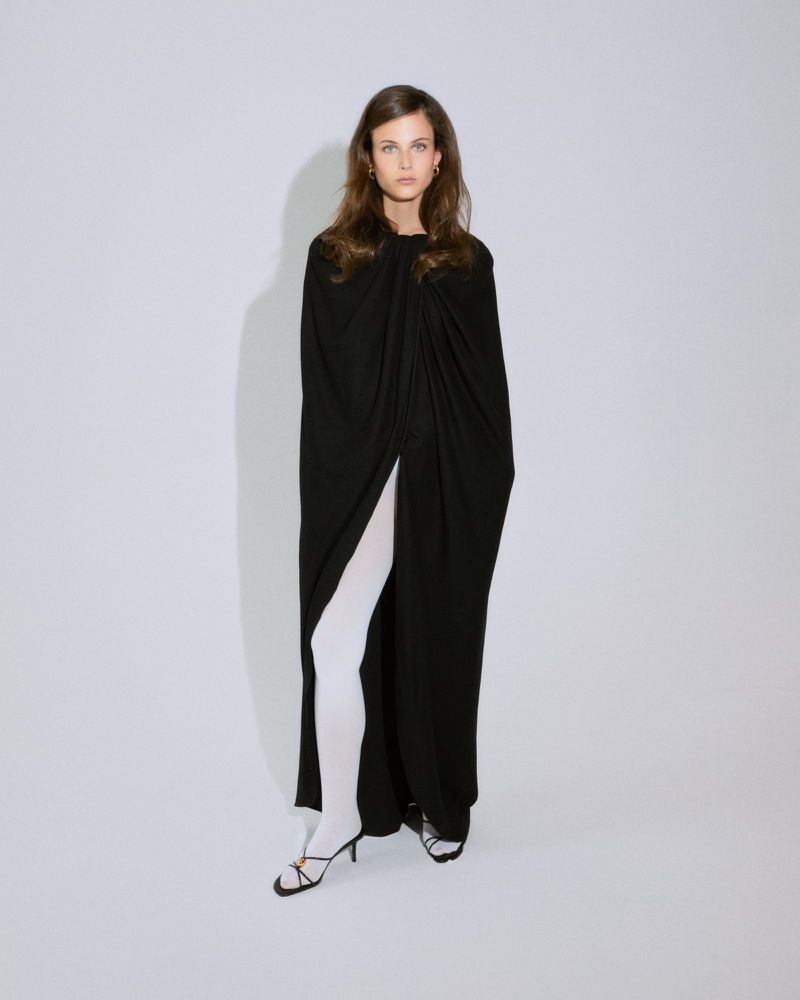 Model standing against a studio backdrop wearing a long black draped cape, opaque white tights, and strappy black heels.