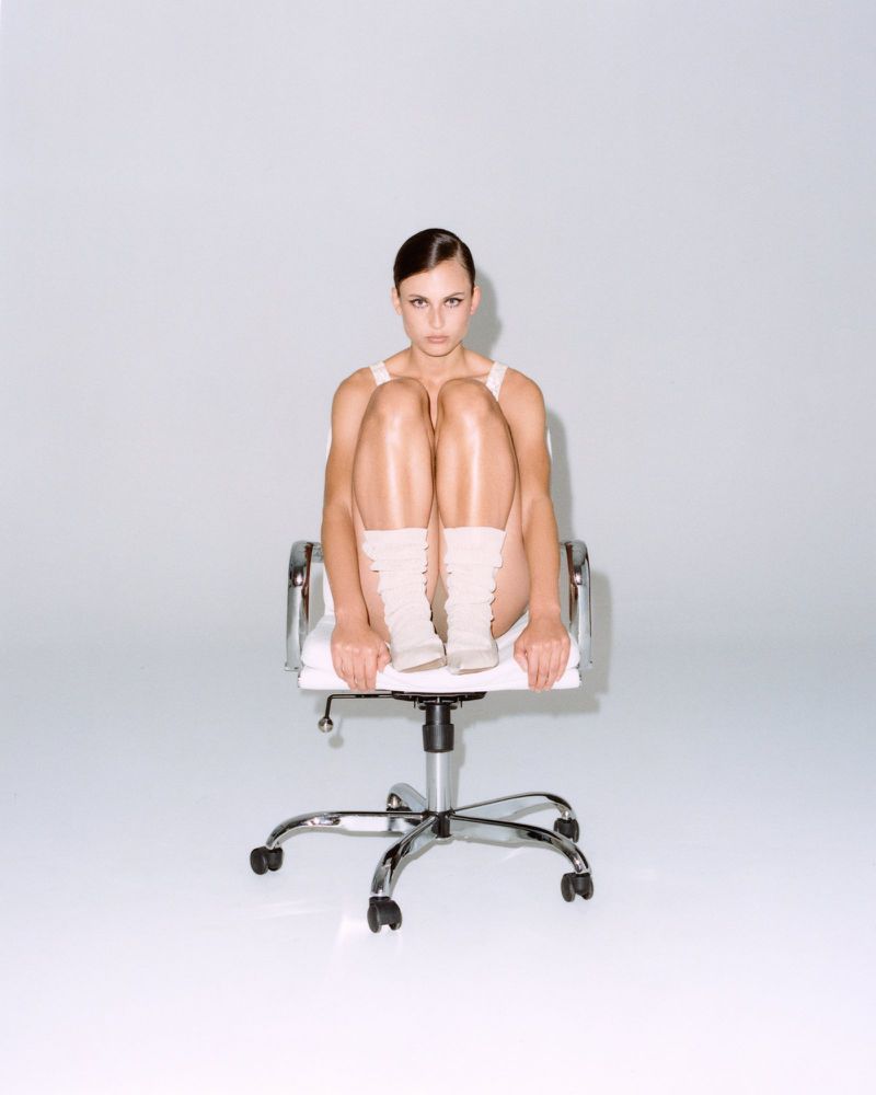 An adult woman sits in an office chair with her knees up to her chest. The composition focuses on her against a neutral backdrop.