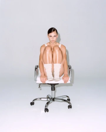 An adult woman sits in an office chair with her knees up to her chest. The composition focuses on her against a neutral backdrop.