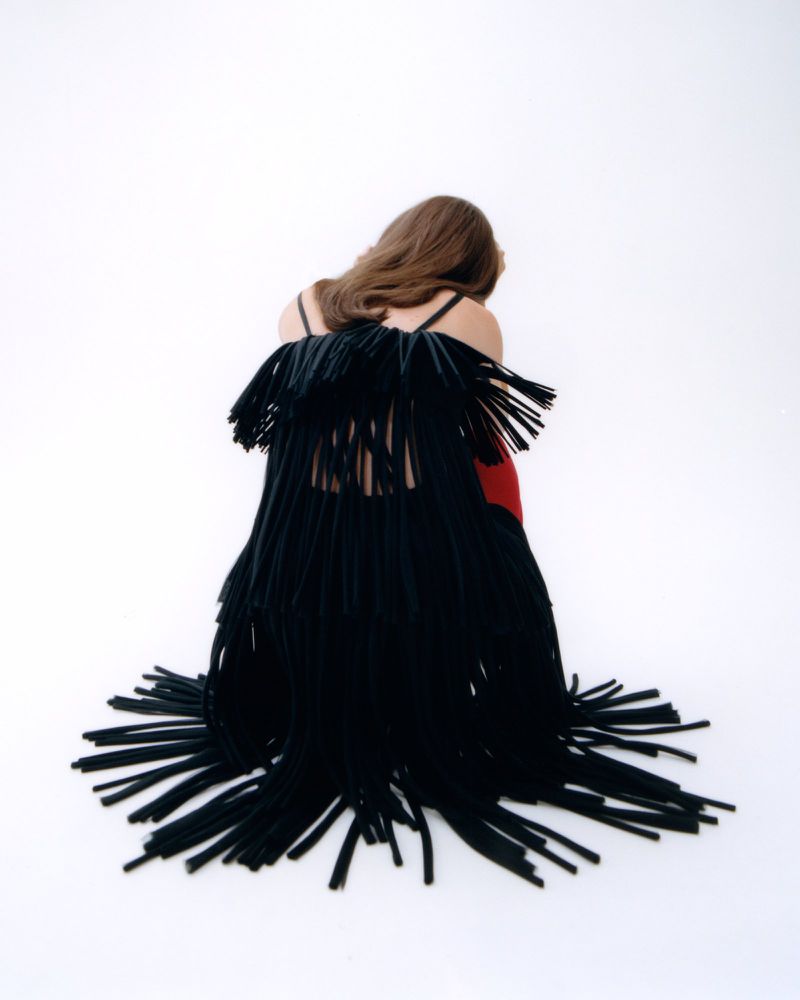 Back view of a person with long brown hair in a black fringe-trimmed garment against a clean white backdrop in a brightly lit photo.