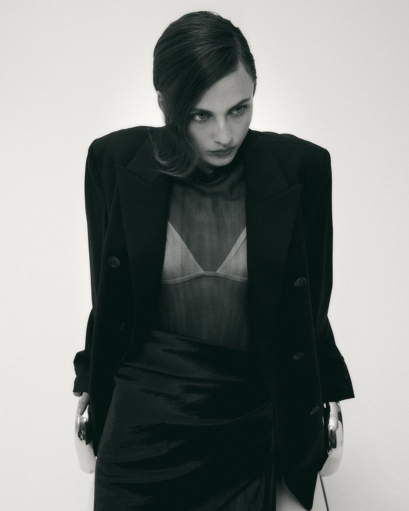 Model wearing an oversized black blazer and sheer mesh top seated on a modern chair against a bright white background.