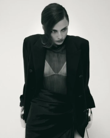 Model wearing an oversized black blazer and sheer mesh top seated on a modern chair against a bright white background.