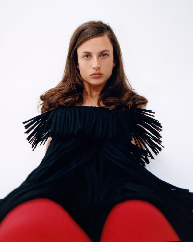Close-up portrait of a young adult woman with long brown hair wearing a black dress with fringe, looking directly at the camera.