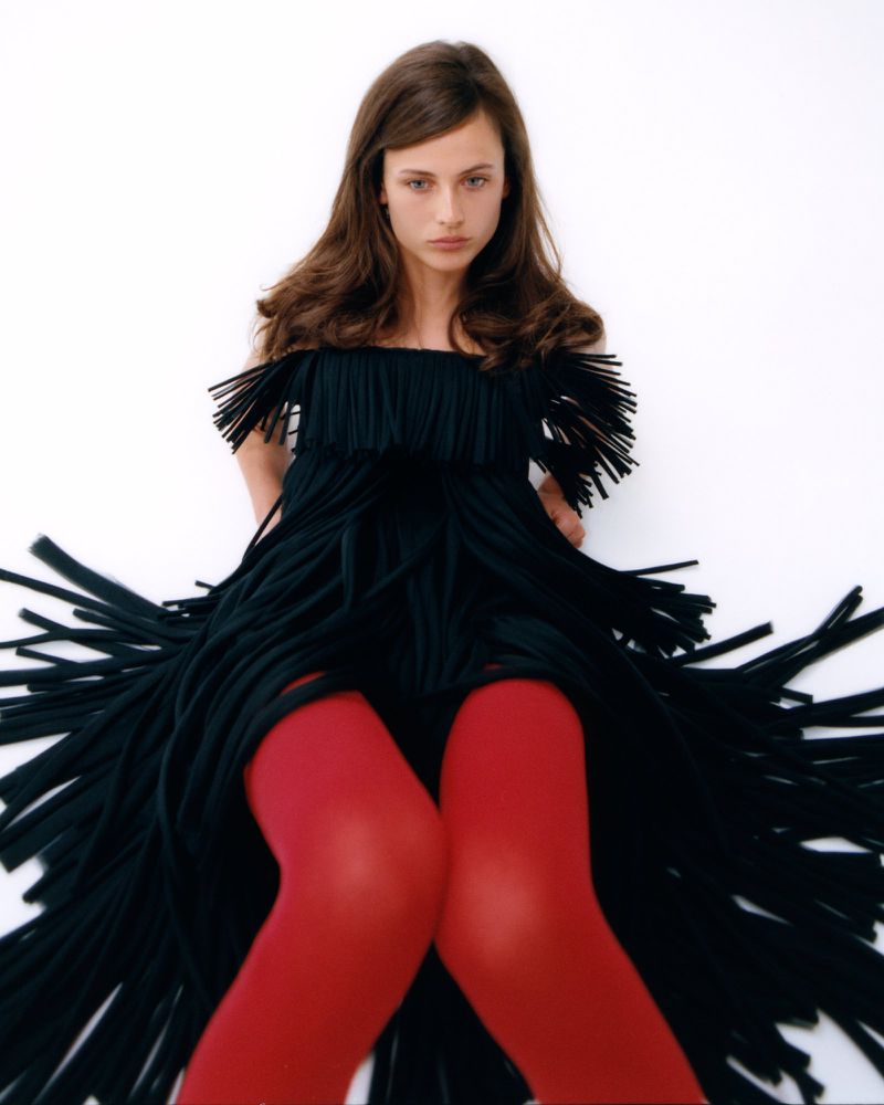 Fashion model in a textured black fringe dress and vibrant red tights seated against a pure white studio background.