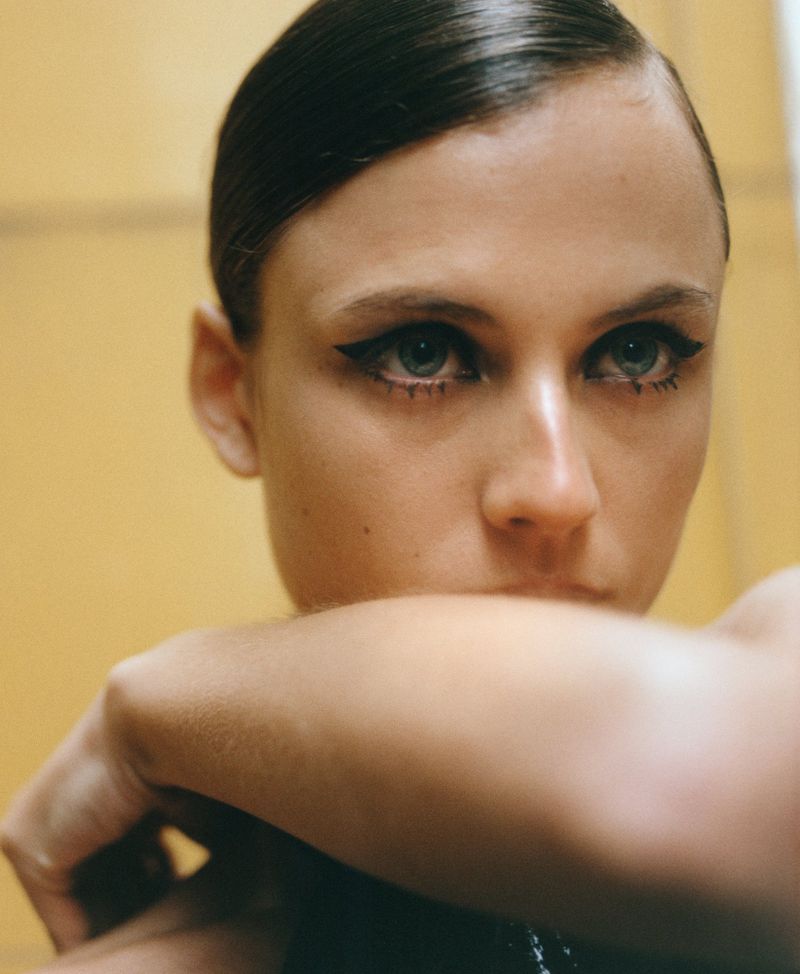 Close-up portrait of an adult female with slicked-back hair, heavy black eyeliner, resting her chin on her arm, against a yellow background.