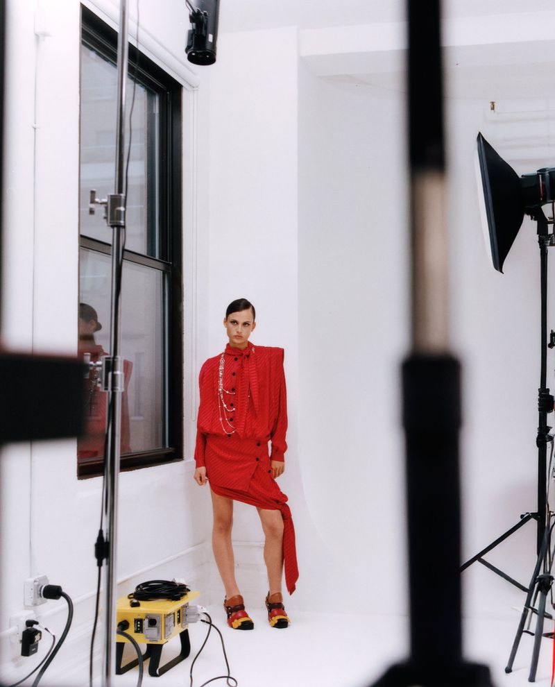 Adult female fashion model poses in a red dress in a photo studio with lighting equipment and a window. Full shot.