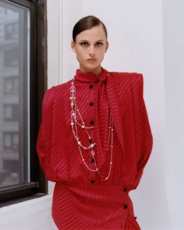 Model with sleek hair wearing a high-neck red blouse with padded shoulders and layered pearl and silver chain necklaces by a window.