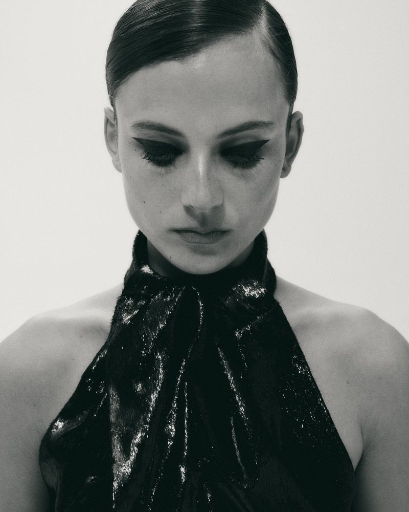 Portrait of an adult female with dark hair and striking winged eyeliner, wearing a black halter dress against a bright white backdrop.