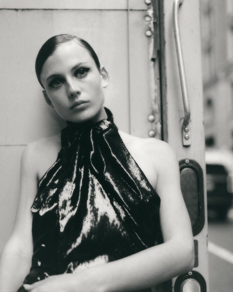 Black and white portrait of a woman in a glossy high-neck halter top leaning against a metal vehicle exterior.