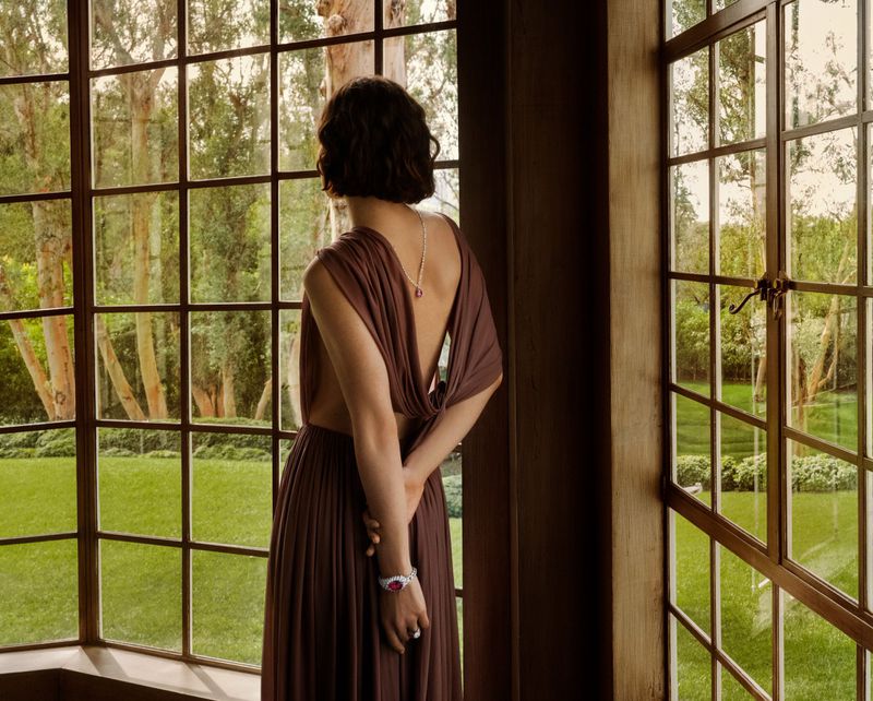 Woman in elegant brown dress gazes through window with green outdoor view. Natural light, back view, jewelry details. Indoor setting.