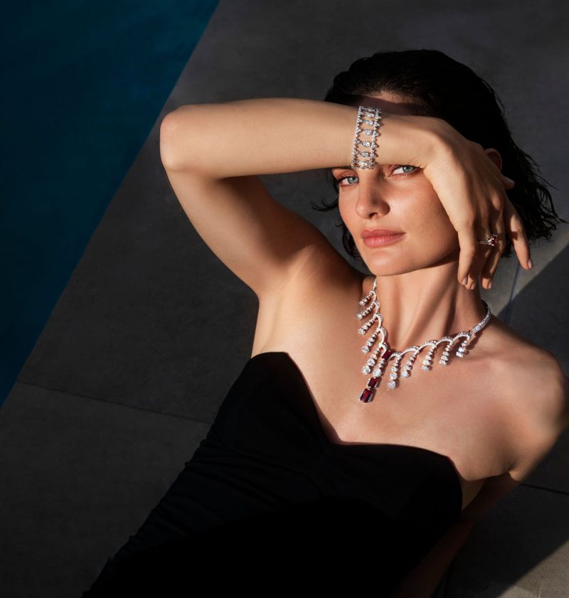 Woman poses wearing white gold and ruby necklace and bracelet in studio with black dress and dramatic lighting near a blue wall.