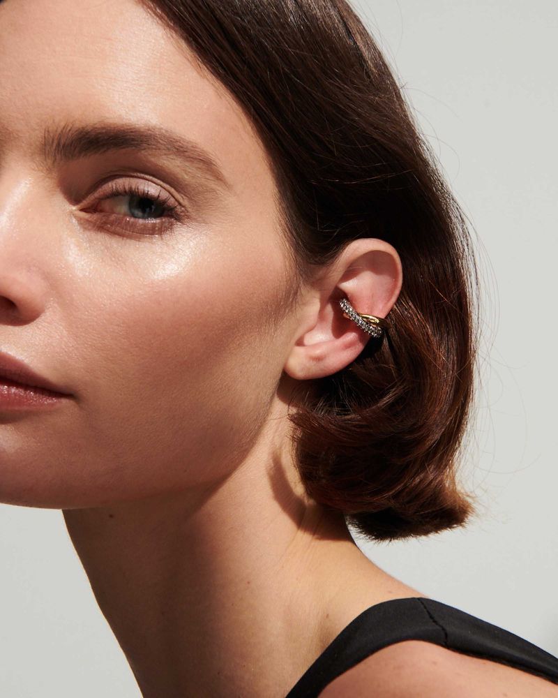 Close-up of a woman's ear adorned with a decorative ear cuff featuring gold and diamond-like accents, set against a soft grey backdrop.