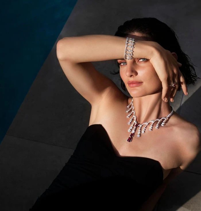 Model Delfina Morbelli wearing a luxury diamond necklace, bracelet, and black strapless dress reclining by a pool in sunlight.