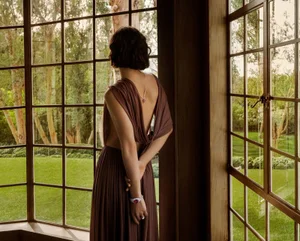 A woman in a brown dress standing by a large grid window looking out at a green garden, captured for Cartier.
