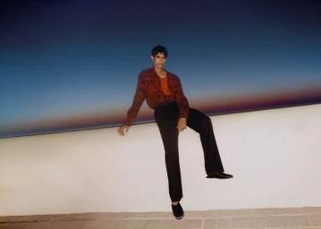 Man wearing a patterned red shirt and black trousers sitting on a low wall against a sunset sky.