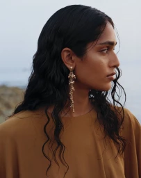 Profile portrait of a South Asian woman wearing long gold statement earrings and a brown top outdoors by the coast.