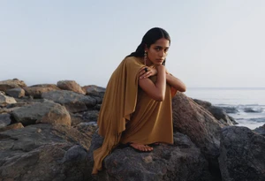 Model in a draped ochre dress crouching on dark rocks near the ocean during the H&M Diwali Collection 2026 campaign shoot.