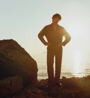Model standing on rocky shore against sunset sky and ocean background, wearing dark collared shirt and trousers with hands on hips.