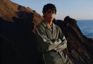 A young male model with dark hair wearing an olive green collared shirt standing against rocky terrain near the sea at sunset.