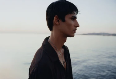 Profile portrait of a young man wearing a dark shirt and silver necklace by the ocean at dusk.
