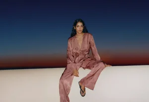 Model seated on a white wall wearing a dusty rose silk patterned pantsuit against a dusk sky.