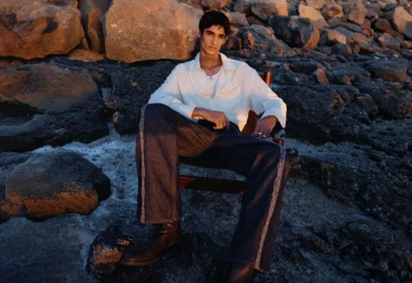Model seated on a wooden chair on a rocky coastal shore wearing a white shirt and dark trousers at sunset.