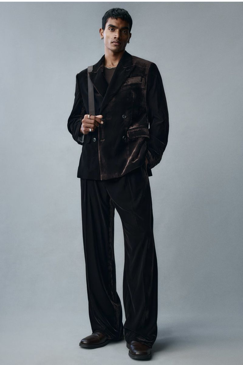 Man wearing a dark velvet double-breasted suit and trousers against a neutral grey studio background