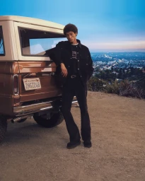 Model leaning against a vintage brown car on a dusty hilltop overlooking a city at dusk for Amiri