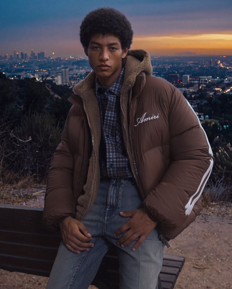 Model in brown puffer jacket sitting on bench outdoors with city skyline background at dusk