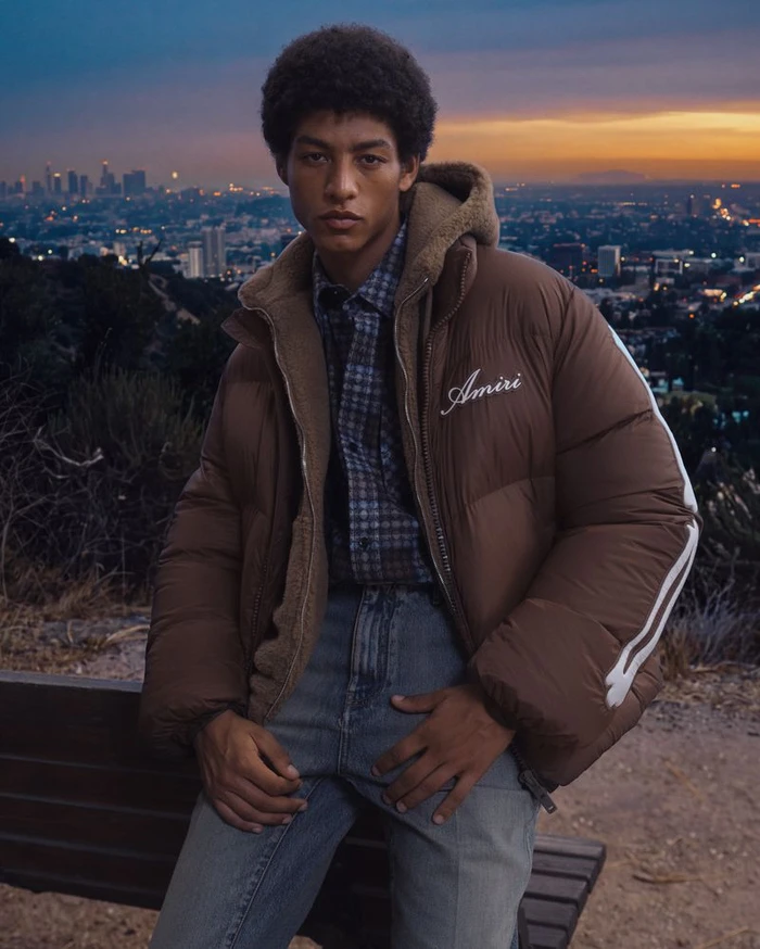 Model in brown puffer jacket sitting on bench outdoors with city skyline background at dusk