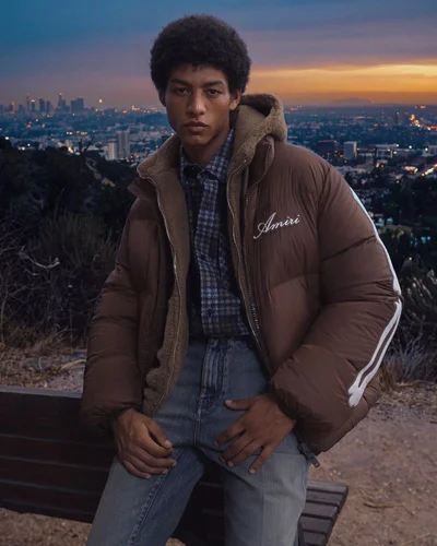 Model in brown puffer jacket sitting on bench outdoors with city skyline background at dusk