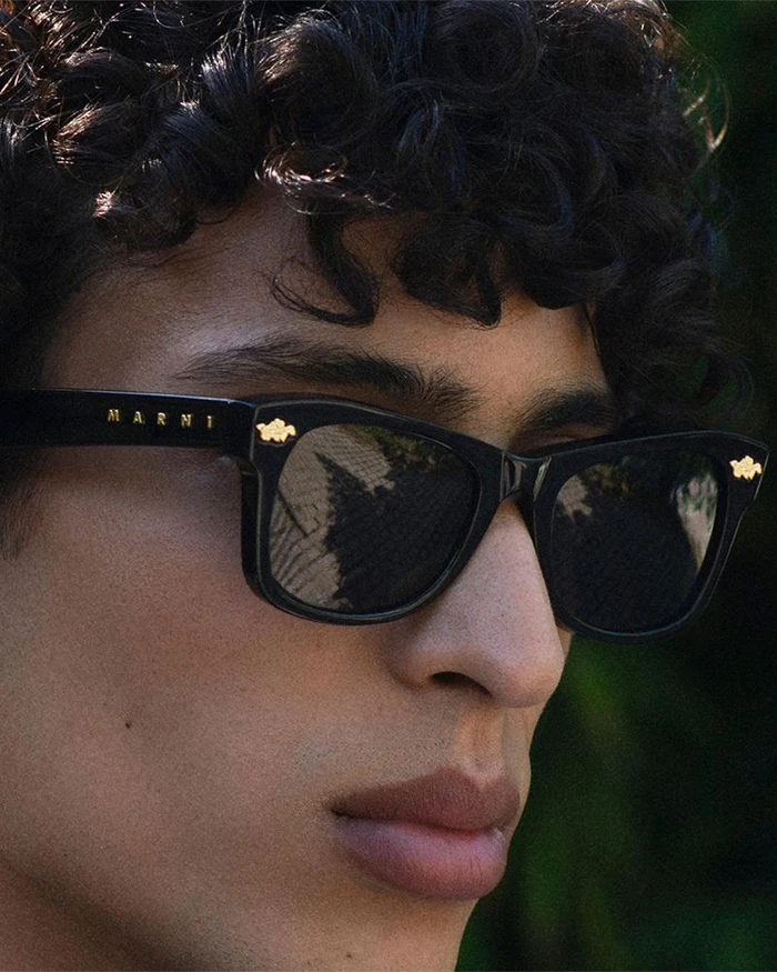 Close-up portrait of a model wearing black Marni sunglasses with gold hardware against an outdoor background