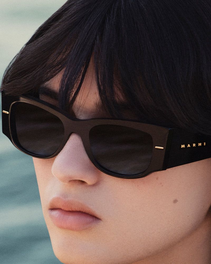 Close-up portrait of a model wearing black rectangular sunglasses with gold branding against an outdoor water background.