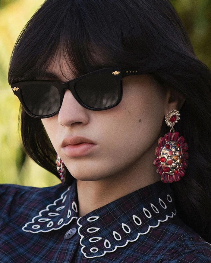 Close-up portrait of a model wearing black sunglasses, statement earrings, and a plaid collared shirt outdoors