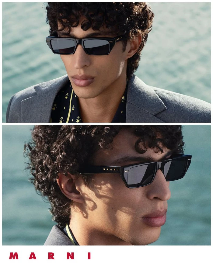 Model wearing black rectangular sunglasses and a grey blazer against an out-of-focus water background for Marni.